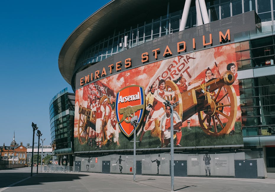 A large, curved glass facade of Emirates Stadium, the home of Arsenal Football Club, featuring a prominent mural that depicts football players dressed in red and white kits celebrating with a vintage 