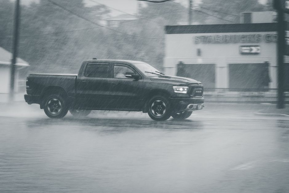 A dark grey pickup truck is driving in heavy rain on a wet urban street, with water spraying from its tires. The vehicle is situated in the foreground, with a modern commercial building featuring a pa